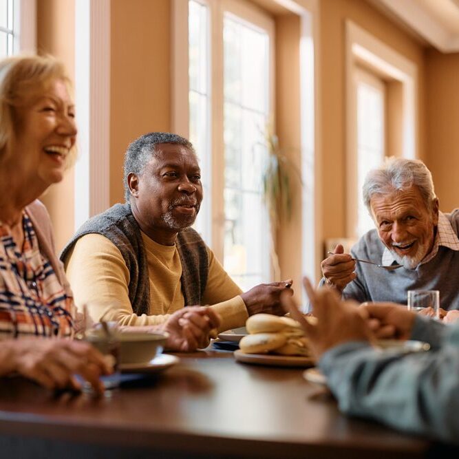 Happy African American senior man talking to his friends while eating at dining table at nursing home.